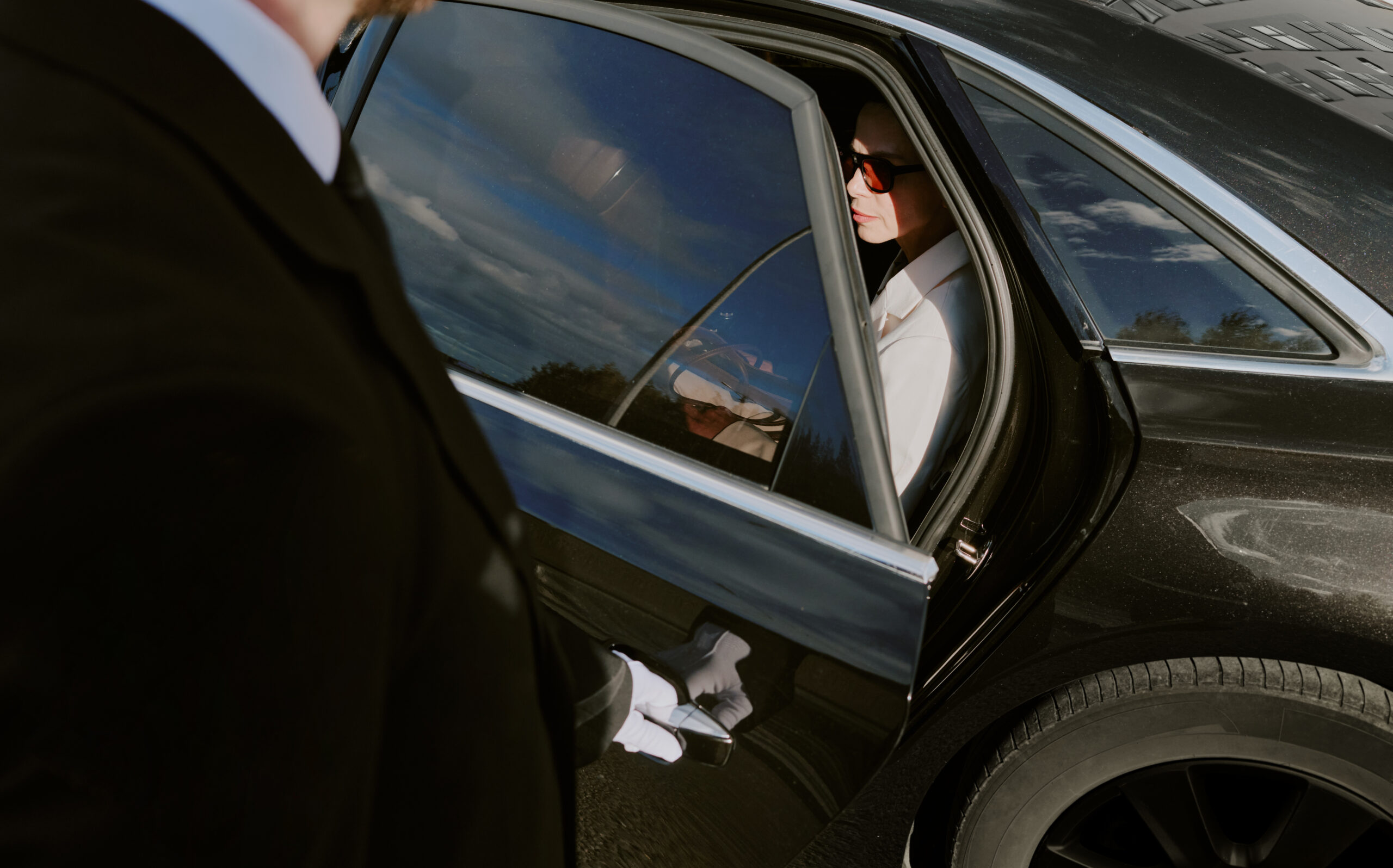 Caucasian young adult man wearing sunglasses sitting in back seat of luxury car while chauffeur in suit and gloves holding door open, business travel concept, outdoor setting